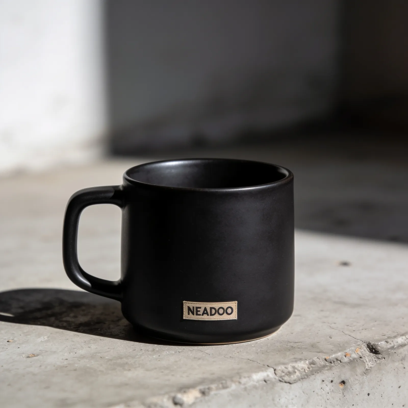 Matte black ceramic mug generated by Seedream 4.5: beige fabric patch with the inscription NEADOO, bright sunlight, concrete windowsill